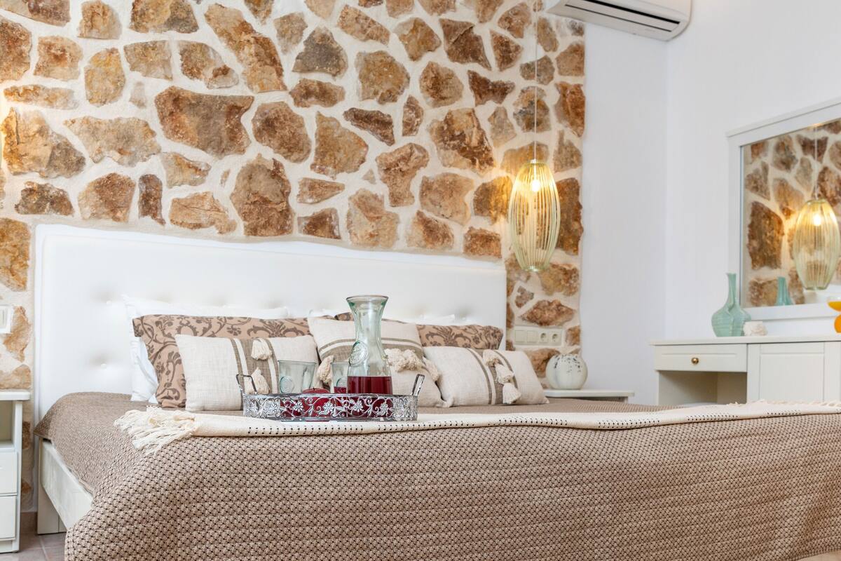 A bedroom with a stone wall, a bed with a patterned headboard, bedside tables, and a dresser with a mirror.