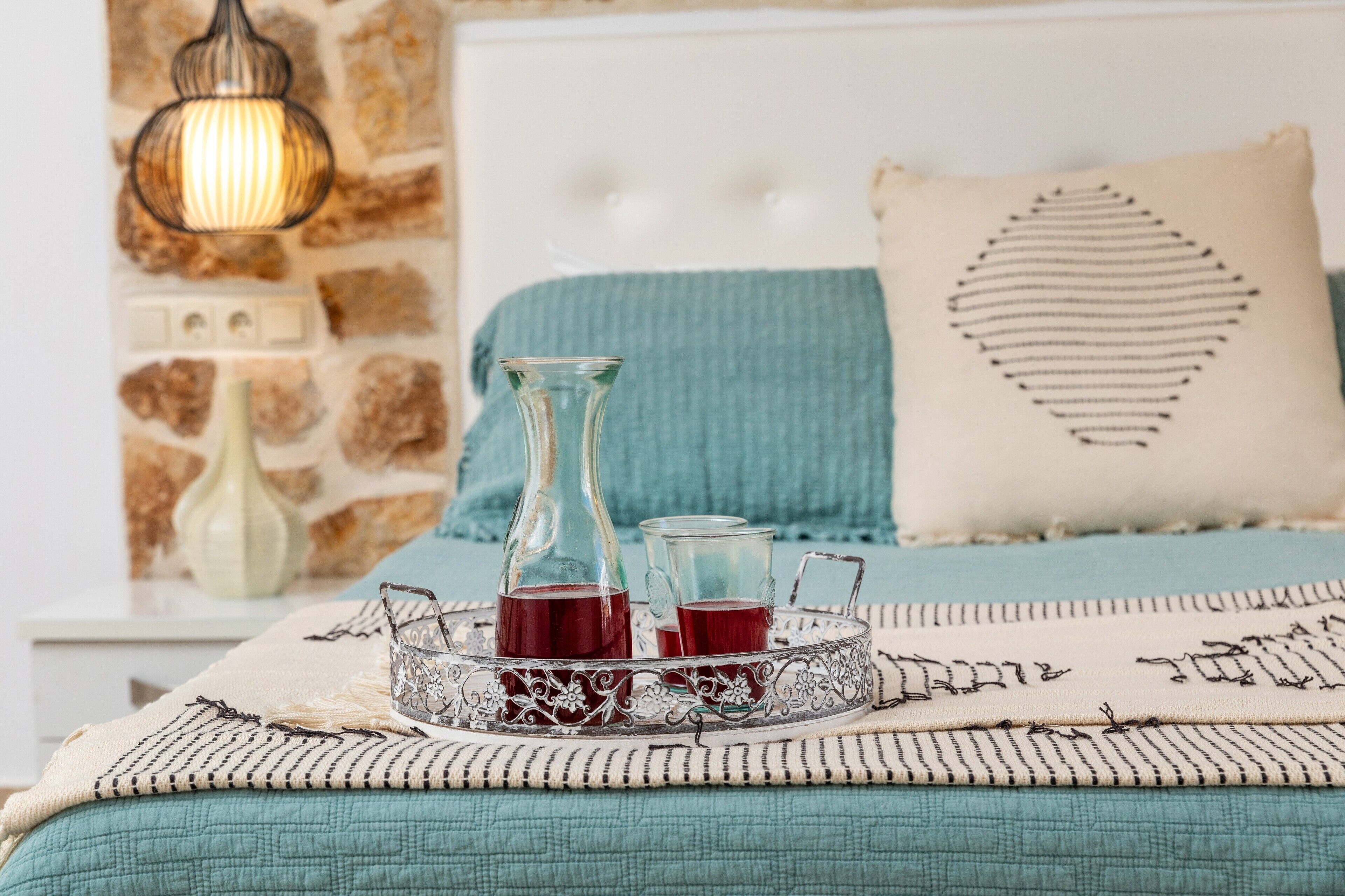 A bed with a teal blanket, a decorative pillow, and a tray with two glasses of red drink.