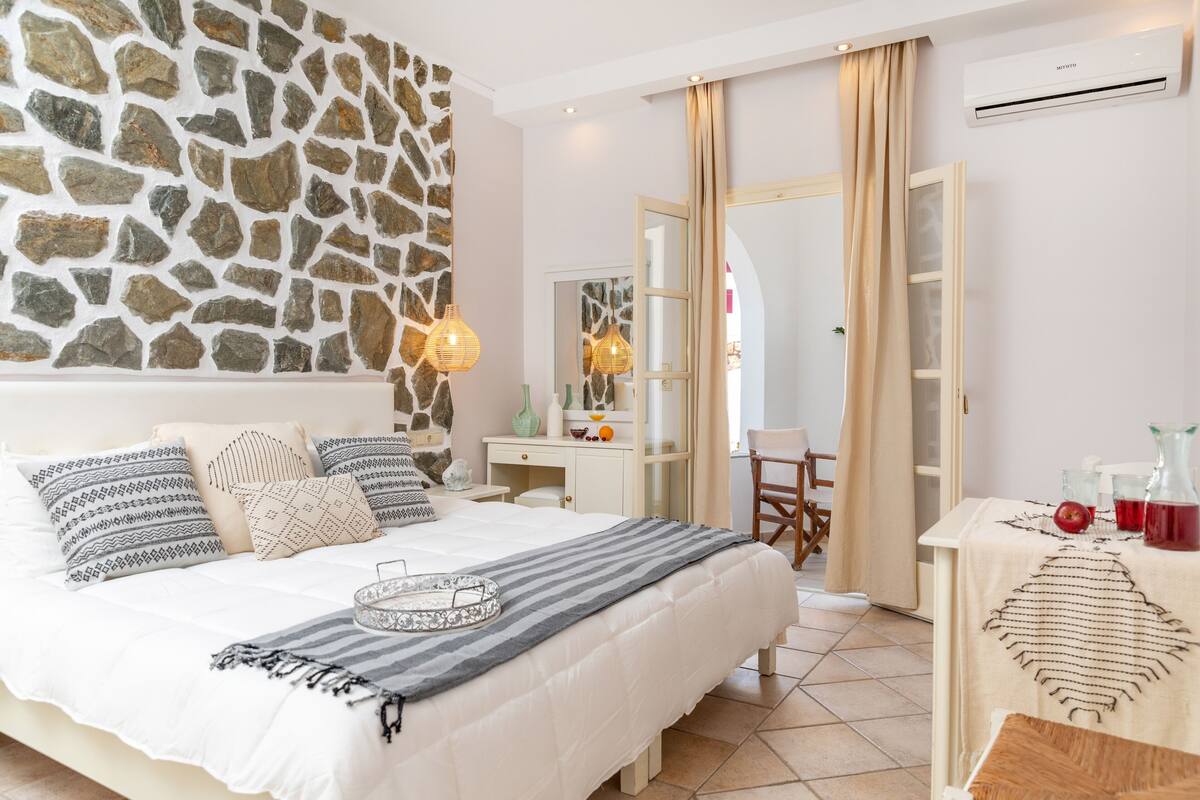 A bedroom with a stone wall, a bed with patterned pillows, a bedside table with a vase, and a view of the bathroom.