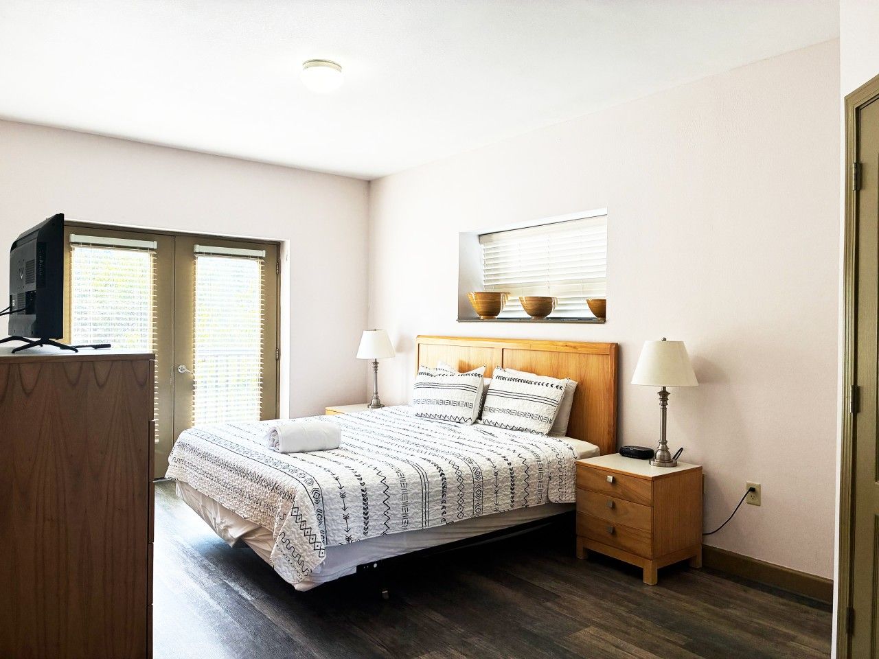 Un dormitorio con cama, mesitas de noche, televisor y ventana con persianas.