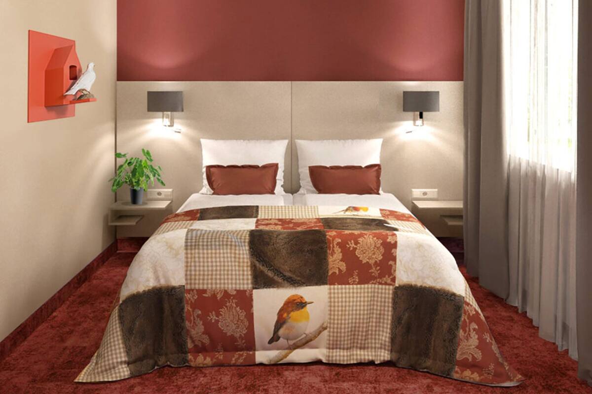 A double bed with a patchwork quilt, two bedside tables with lamps, a red accent wall, and a window with curtains.