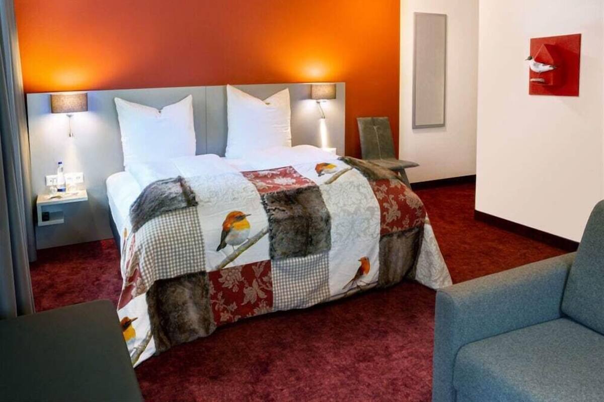 A hotel room with a bed, two bedside lamps, a red carpet, and a blue armchair.