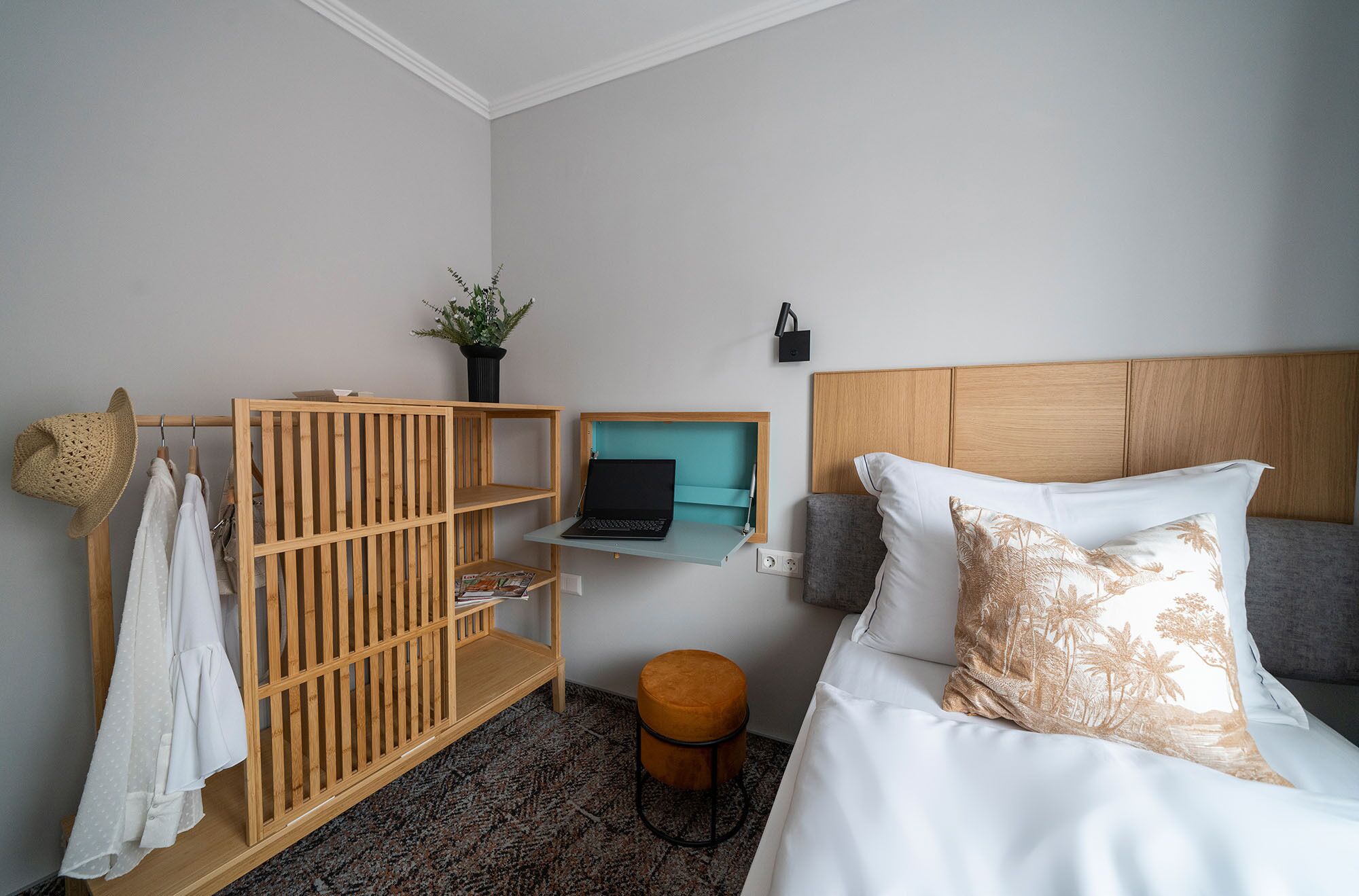 Un dormitorio con una cama, un armario de madera, un taburete pequeño, una computadora portátil sobre una estantería y una planta.