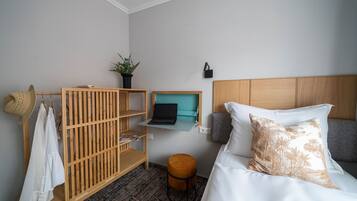 Un dormitorio con una cama, un armario de madera, un taburete pequeño, una computadora portátil sobre una estantería y una planta.