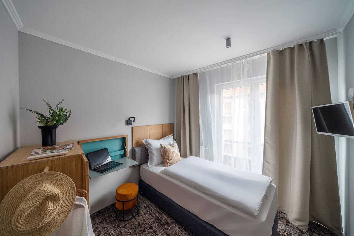 Habitación de hotel con una cama, un escritorio con una computadora portátil, un taburete pequeño, una planta en maceta y un televisor.