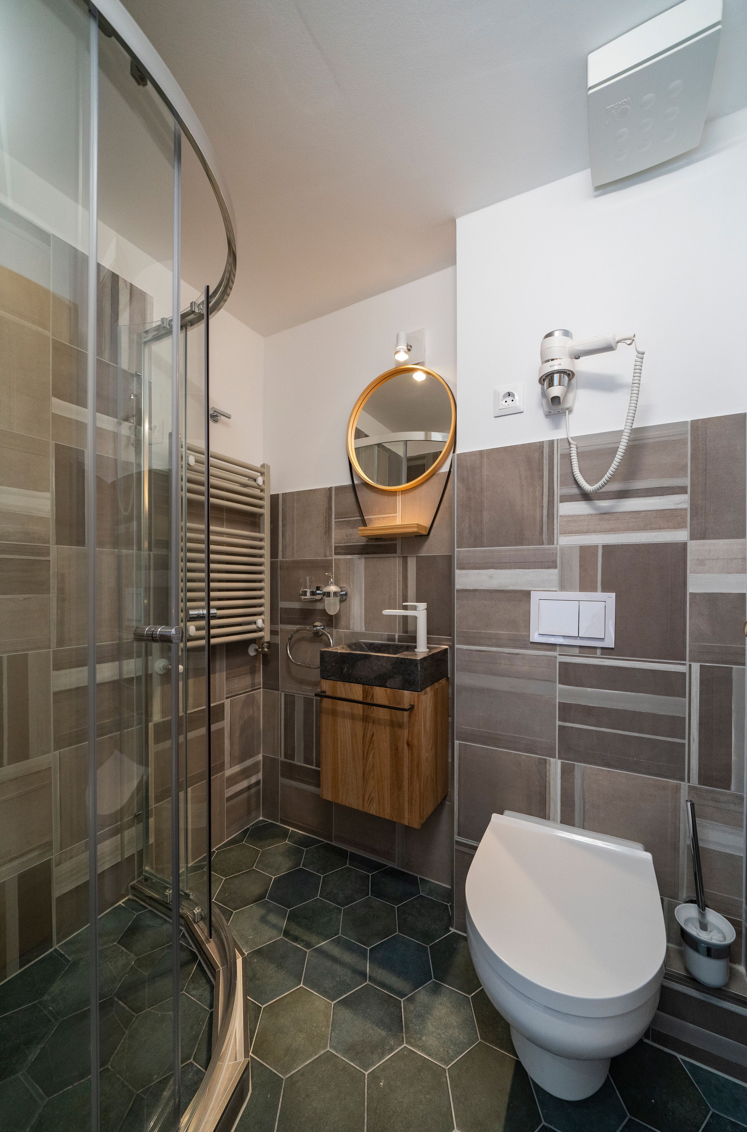Un baño moderno con cabina de ducha de vidrio, tocador de madera, inodoro blanco y espejo con marco redondo.