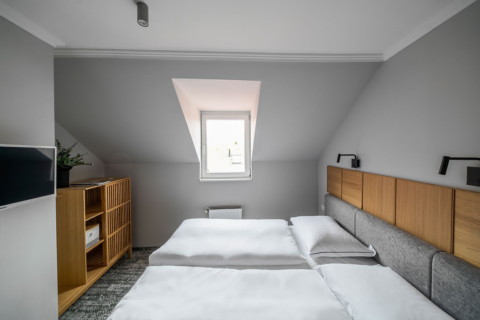 A small, modern bedroom with a sloped ceiling, a skylight, a TV, a wooden wardrobe, and a grey sofa.