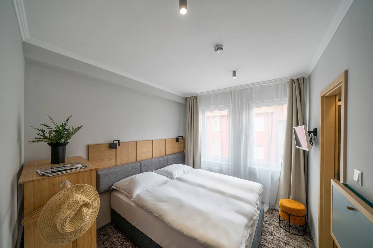 Habitación de hotel con cama, escritorio con planta, taburete pequeño y ventana con cortinas.