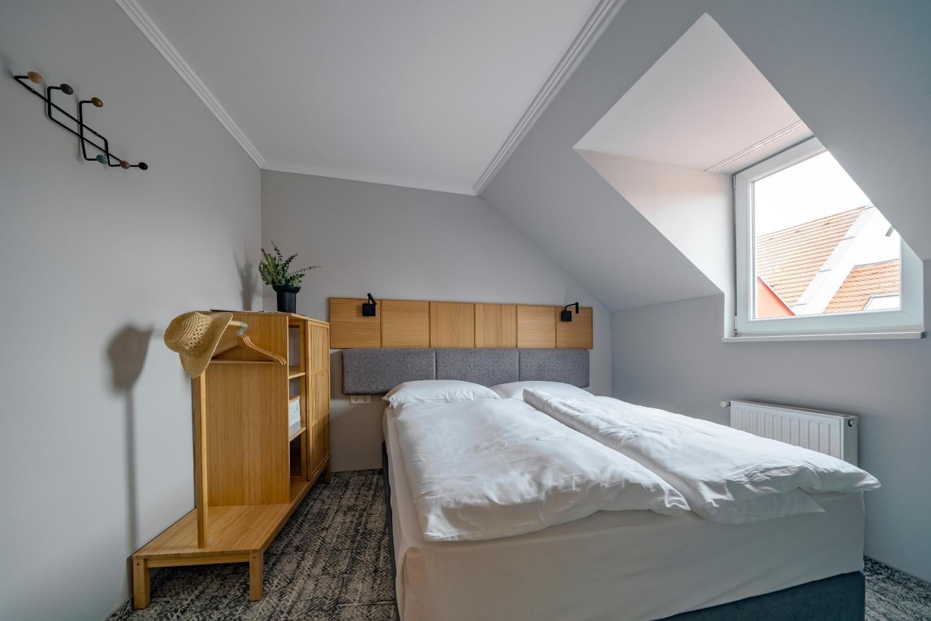 Ein Schlafzimmer mit geneigter Decke, einem Holzschrank, einem Bett mit weißer Bettwäsche, einer Topfpflanze und einem Fenster mit Blick auf Gebäude.