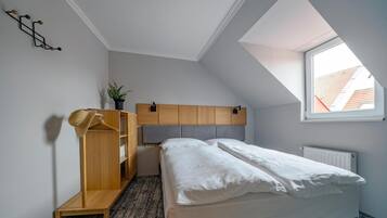 Ein Schlafzimmer mit geneigter Decke, einem Holzschrank, einem Bett mit weißer Bettwäsche, einer Topfpflanze und einem Fenster mit Blick auf Gebäude.