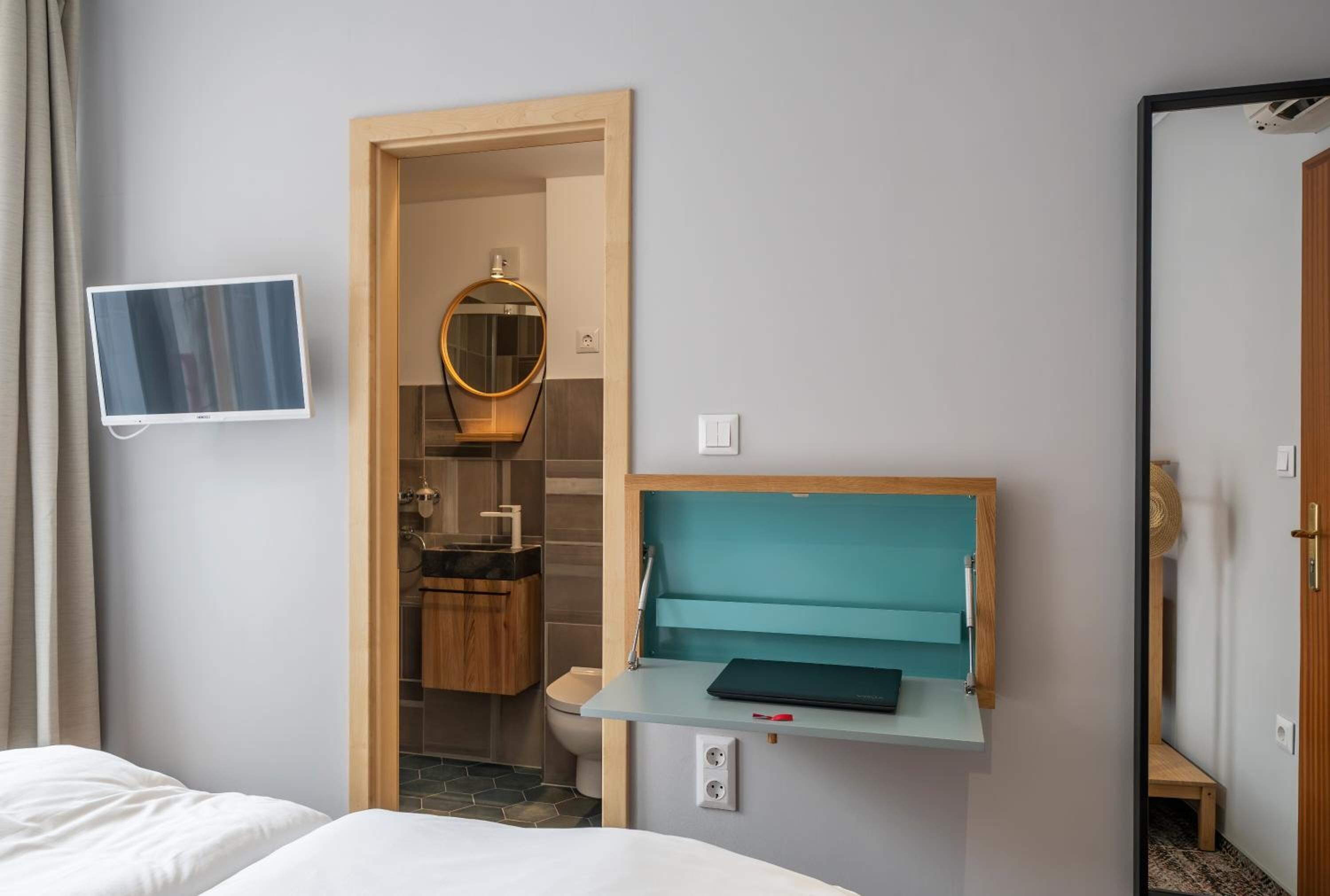 A hotel room with a bed, a desk with a laptop, a mirror, a TV, and a bathroom visible through an open door.