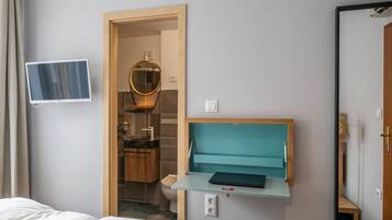 A hotel room with a bed, a desk with a laptop, a mirror, a TV, and a bathroom visible through an open door.