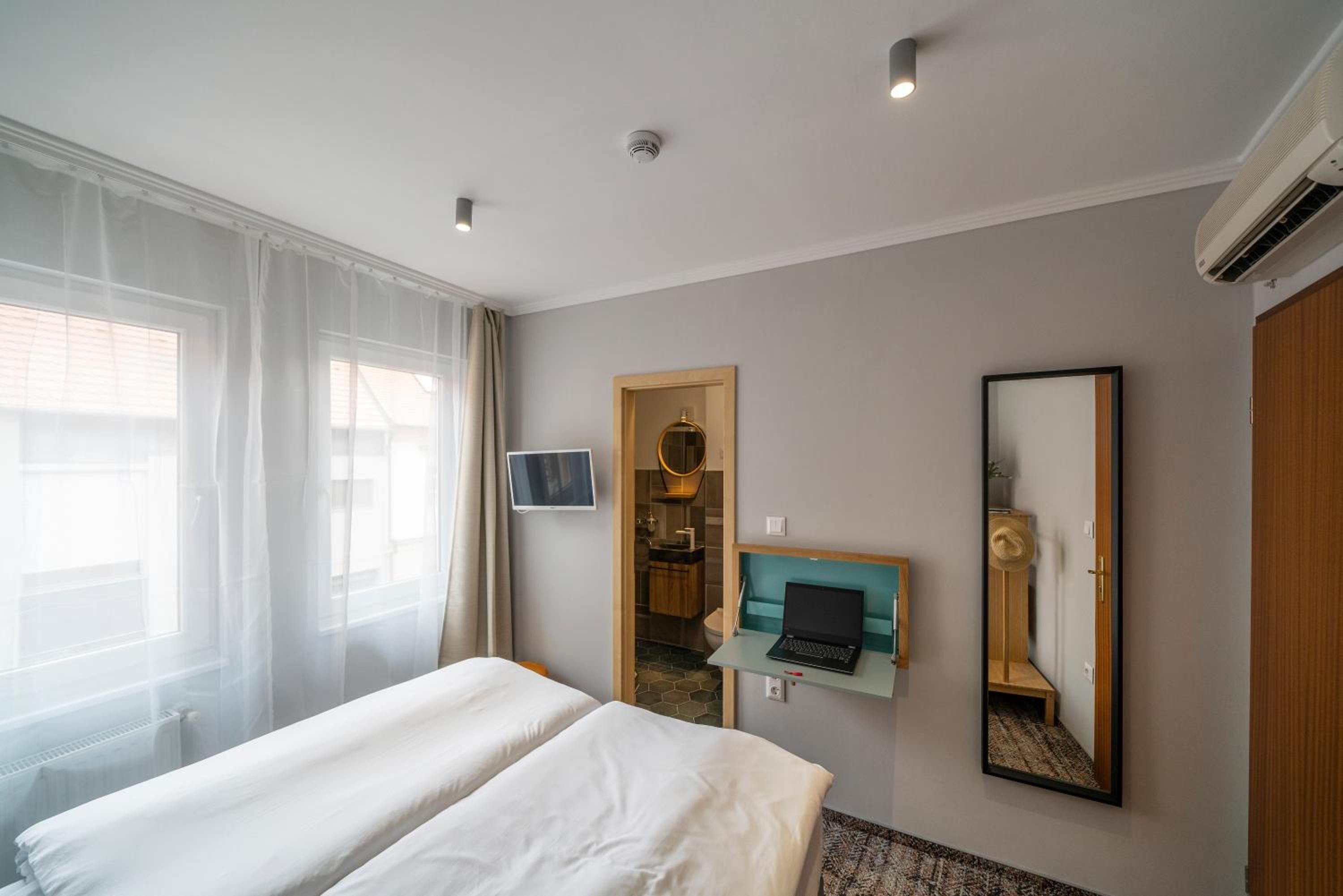 A hotel room with a bed, a desk with a laptop, a mirror, and a bathroom visible through an open door.