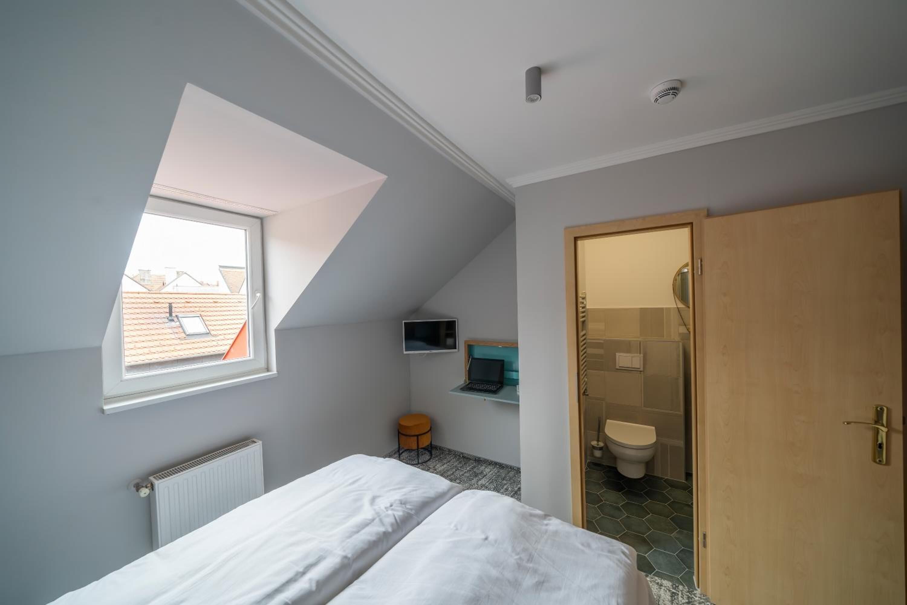 Une chambre avec un puits de lumière, un lit, un bureau avec un ordinateur portable, un petit tabouret et une salle de bain avec des toilettes et une douche.