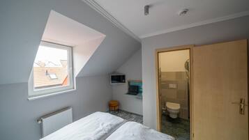 A bedroom with a skylight, a bed, a desk with a laptop, a small stool, and a bathroom with a toilet and shower.