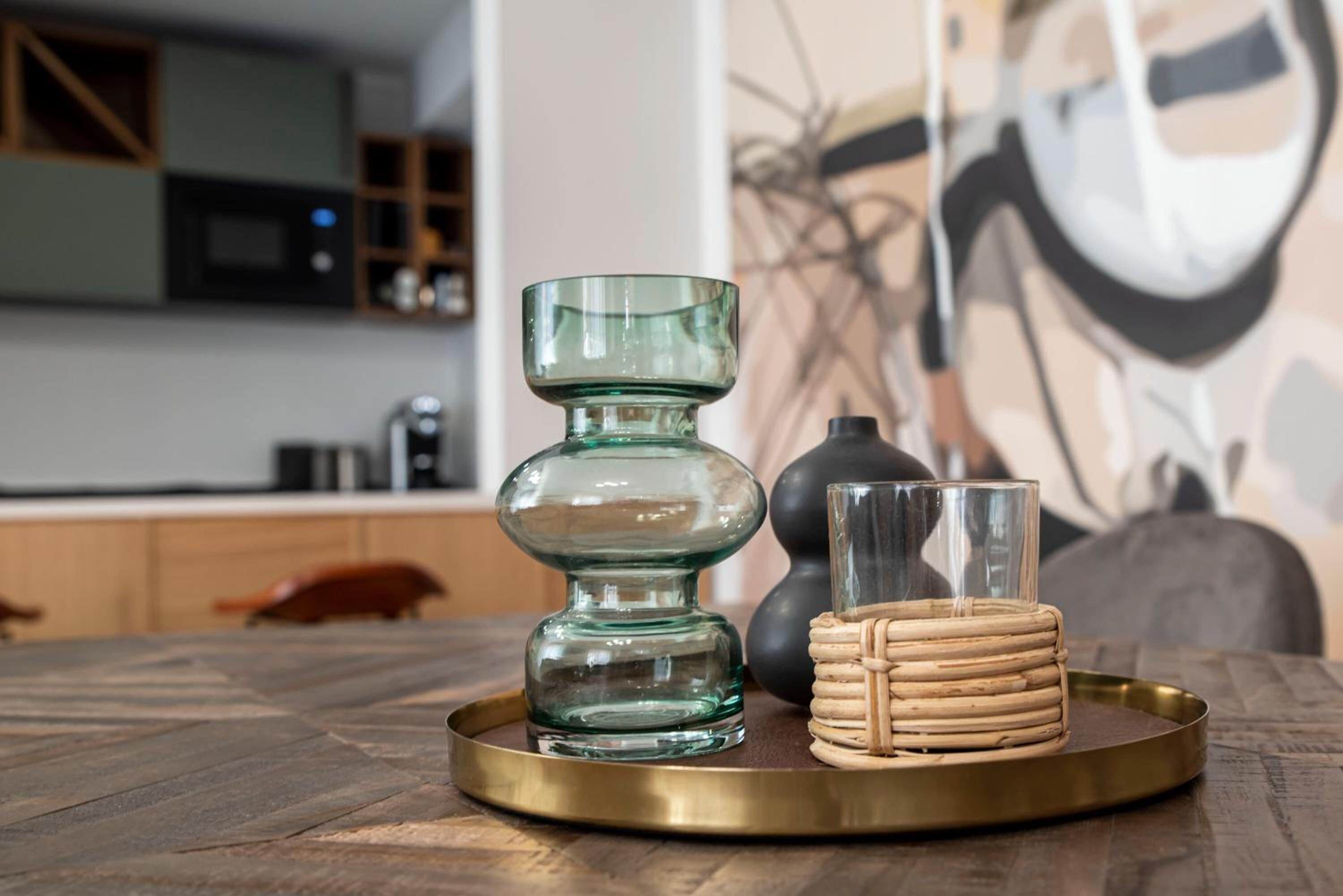 Uma cozinha com uma mesa de madeira, uma tigela de vidro verde, uma pilha de porta-copos e um vaso preto.