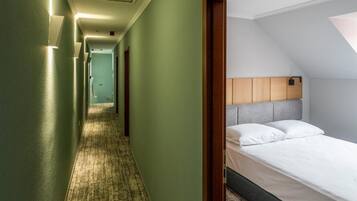 Un pasillo de hotel con pisos alfombrados, una cama con cabecera de madera y una puerta visible al final.