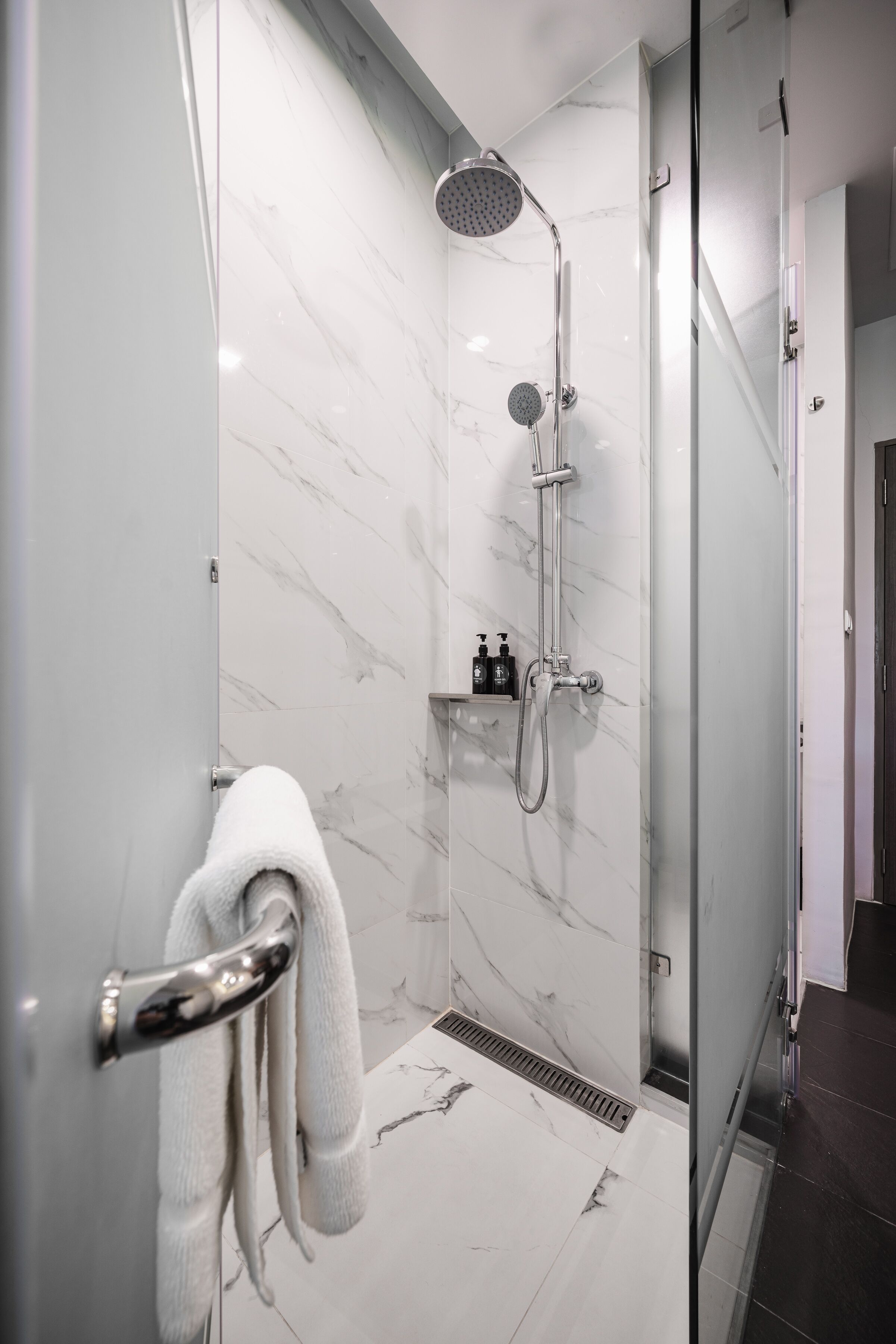 A modern bathroom with a glass shower enclosure, marble walls, a towel rack, and a shelf with toiletries.