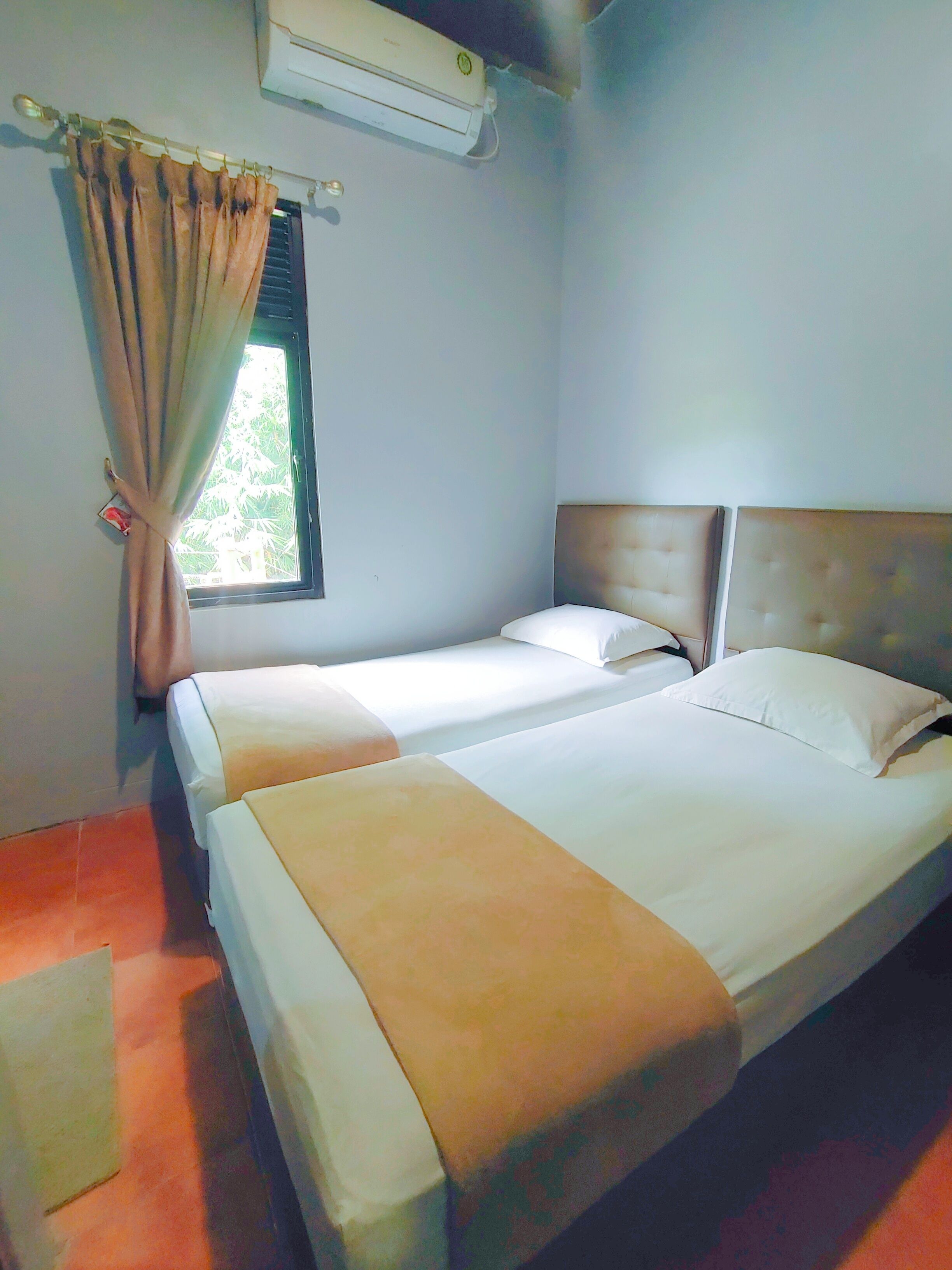 A hotel room with two beds, a window with curtains, an air conditioning unit, and a view of greenery outside.