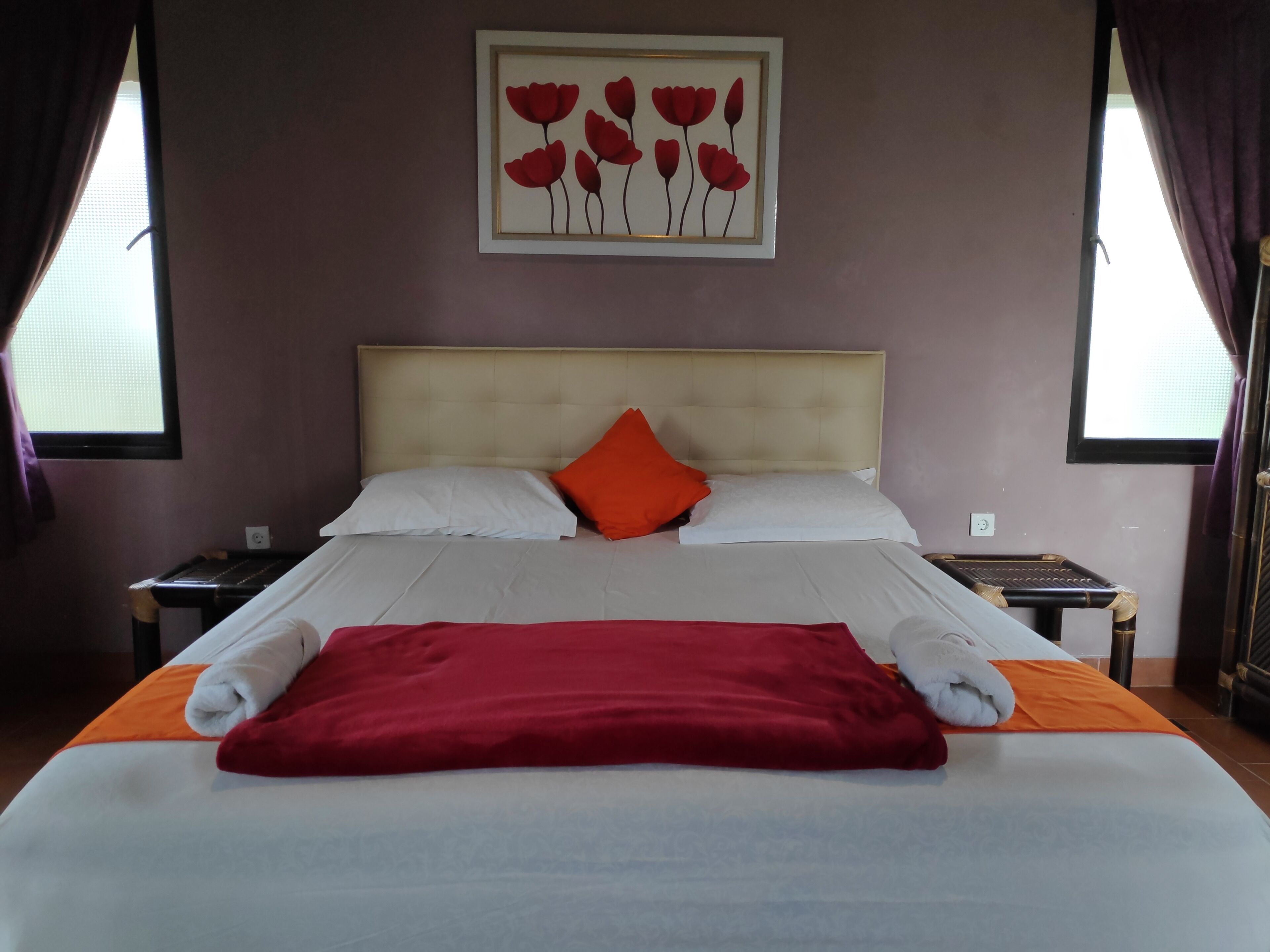 A neatly made bed with white and orange bedding, a red throw blanket, and a framed floral artwork above it.