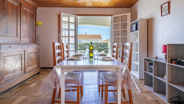 Un coin repas avec une table dressée pour quatre personnes, des meubles en bois et une vue sur la mer par une grande fenêtre.