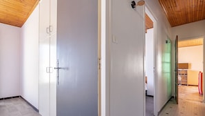 Un couloir aux murs blancs, avec un plafond en bois et un sol carrelé. Il y a des portes sur la gauche et une pièce éclairée de lumière verte sur la droite.