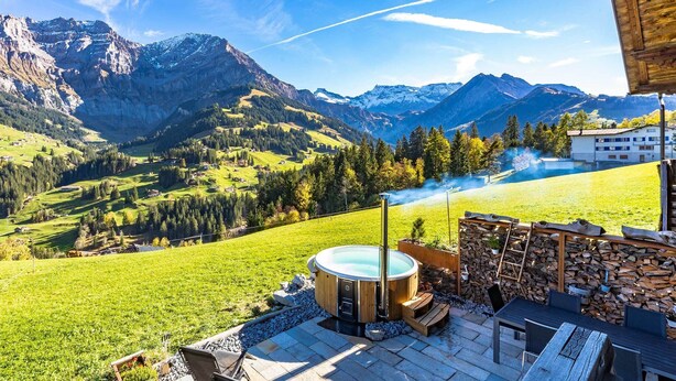 A hot tub on a patio with a mountain view, a wooden deck, and a stone wall.