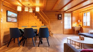A wooden cabin interior with a dining area, a staircase, a clock, and a window with curtains.