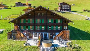 A large wooden building with green shutters, a central entrance, and a small outdoor seating area with a hot tub.