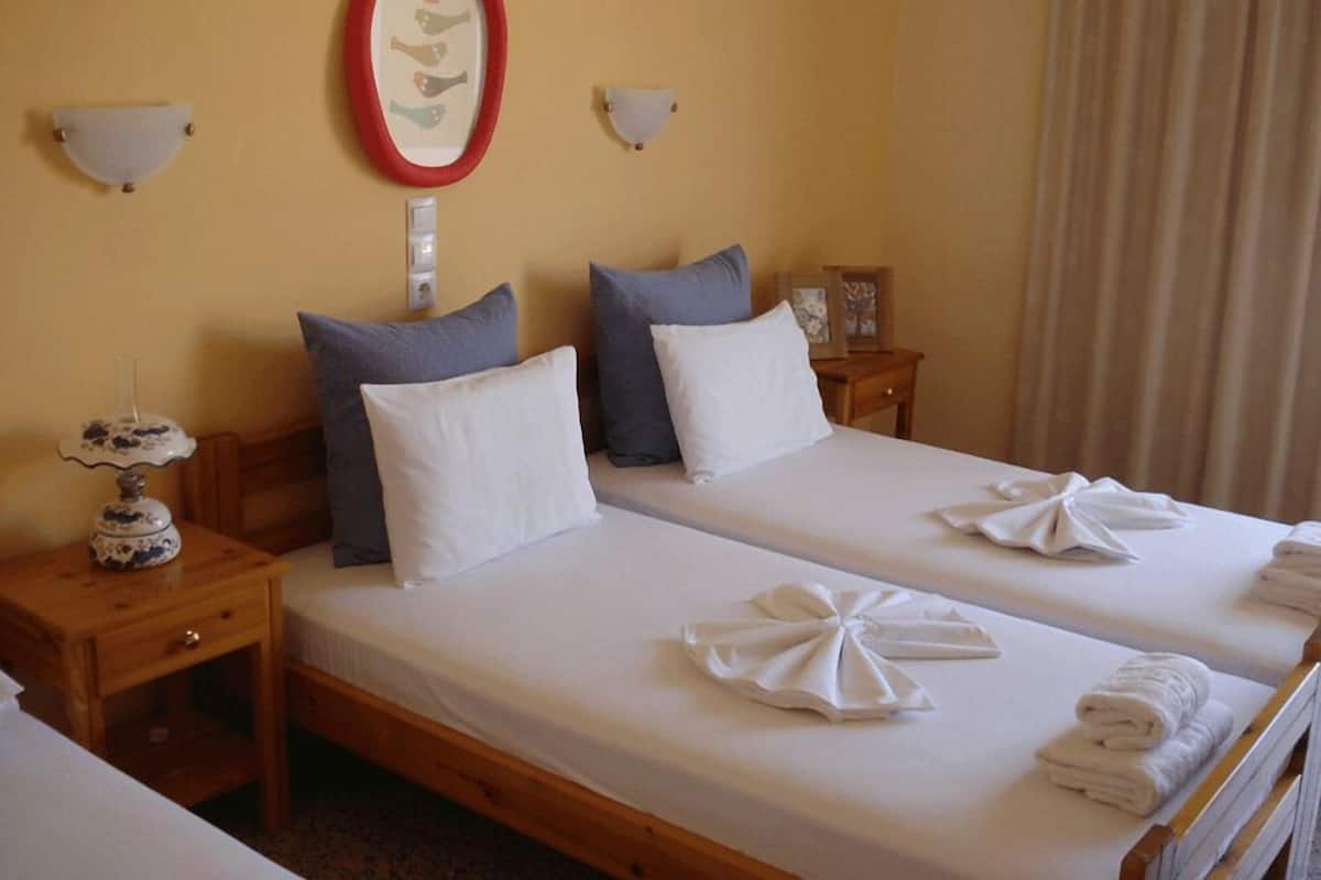 A hotel room with two beds, each with white linens and towels. There are two bedside tables, one with a lamp and the other with a picture frame. The room has a simple, clean design with a neutral color palette.