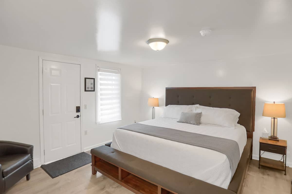 A bedroom with a bed, bedside tables, a bench, a door, and a window with blinds.
