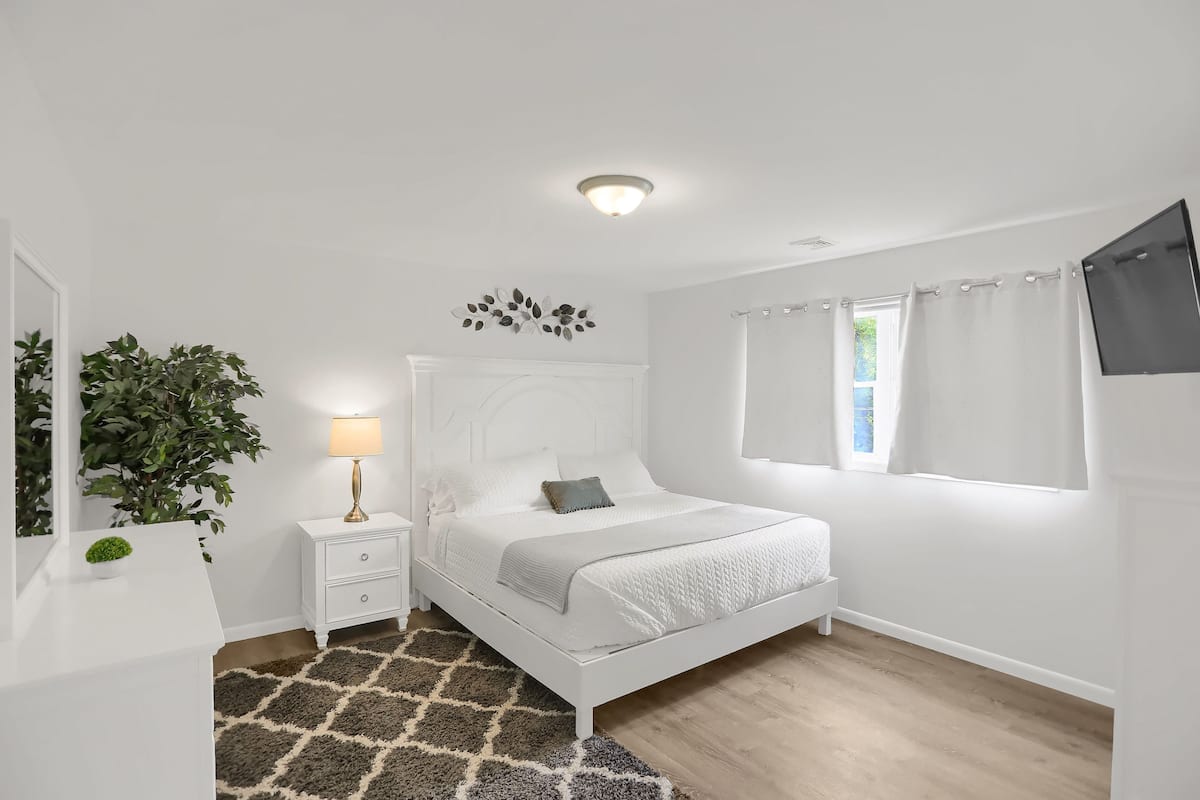A white bedroom with a bed, bedside table, lamp, and a large potted plant.