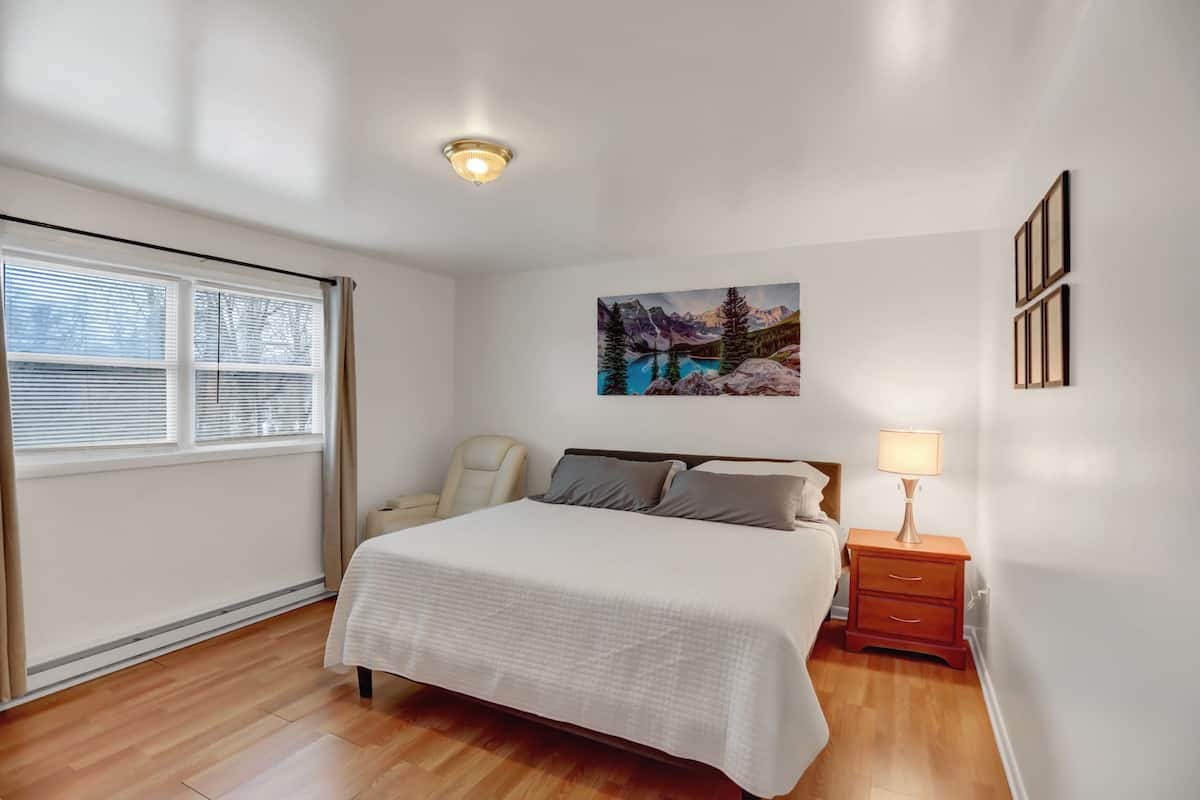 A bedroom with a bed, a chair, a nightstand with a lamp, a window with blinds, and a painting on the wall.