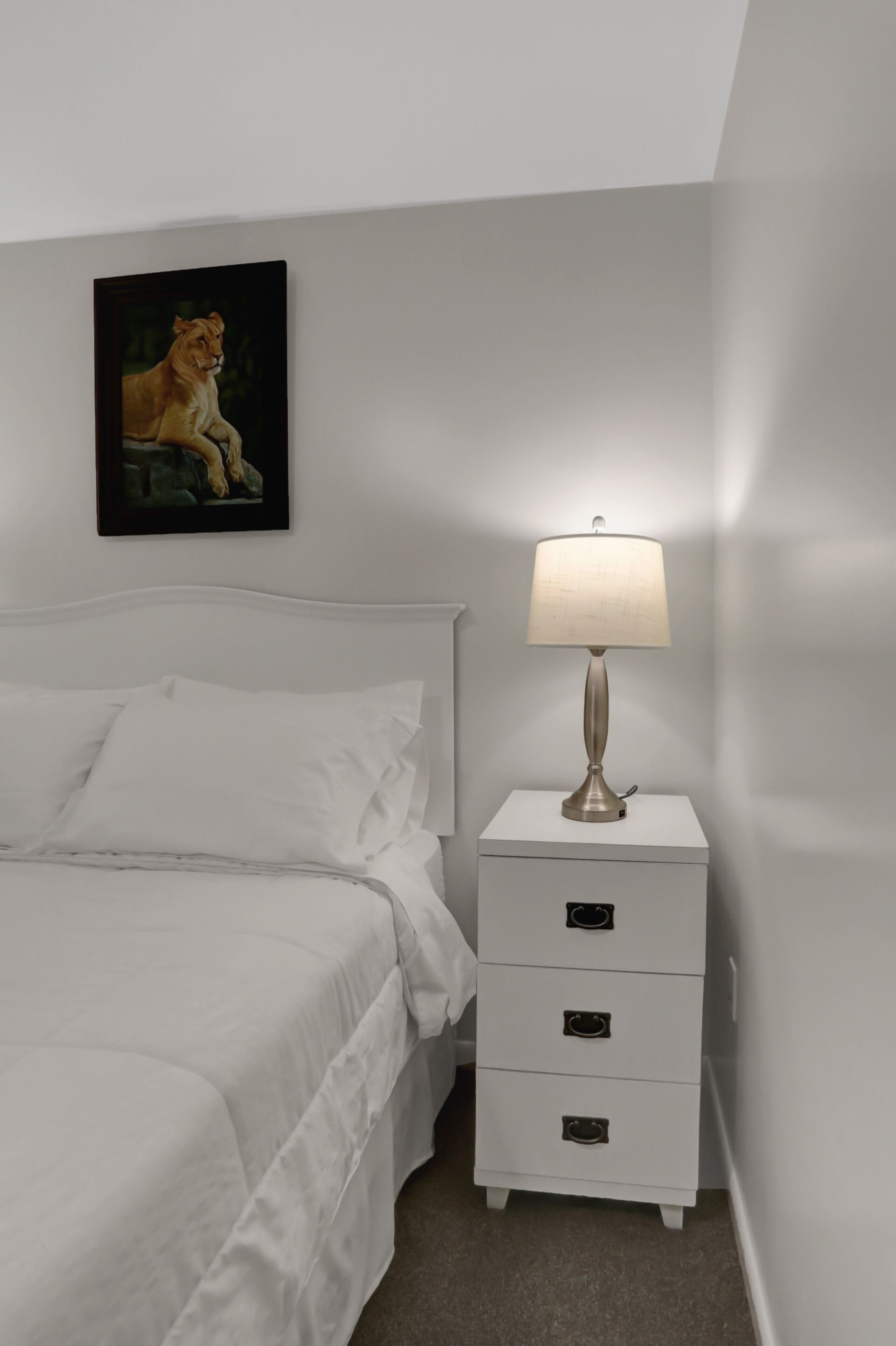 Una cama bien hecha con sábanas blancas, una mesita de noche blanca con cajones, una lámpara fijada a la pared y un cuadro con un león.