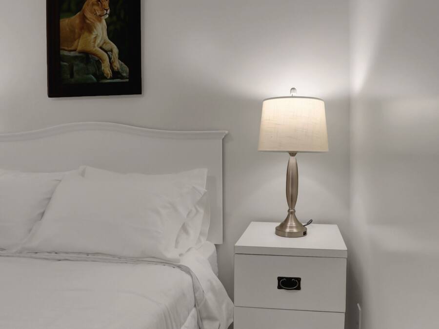 Una cama bien hecha con sábanas blancas, una mesita de noche blanca con cajones, una lámpara fijada a la pared y un cuadro con un león.