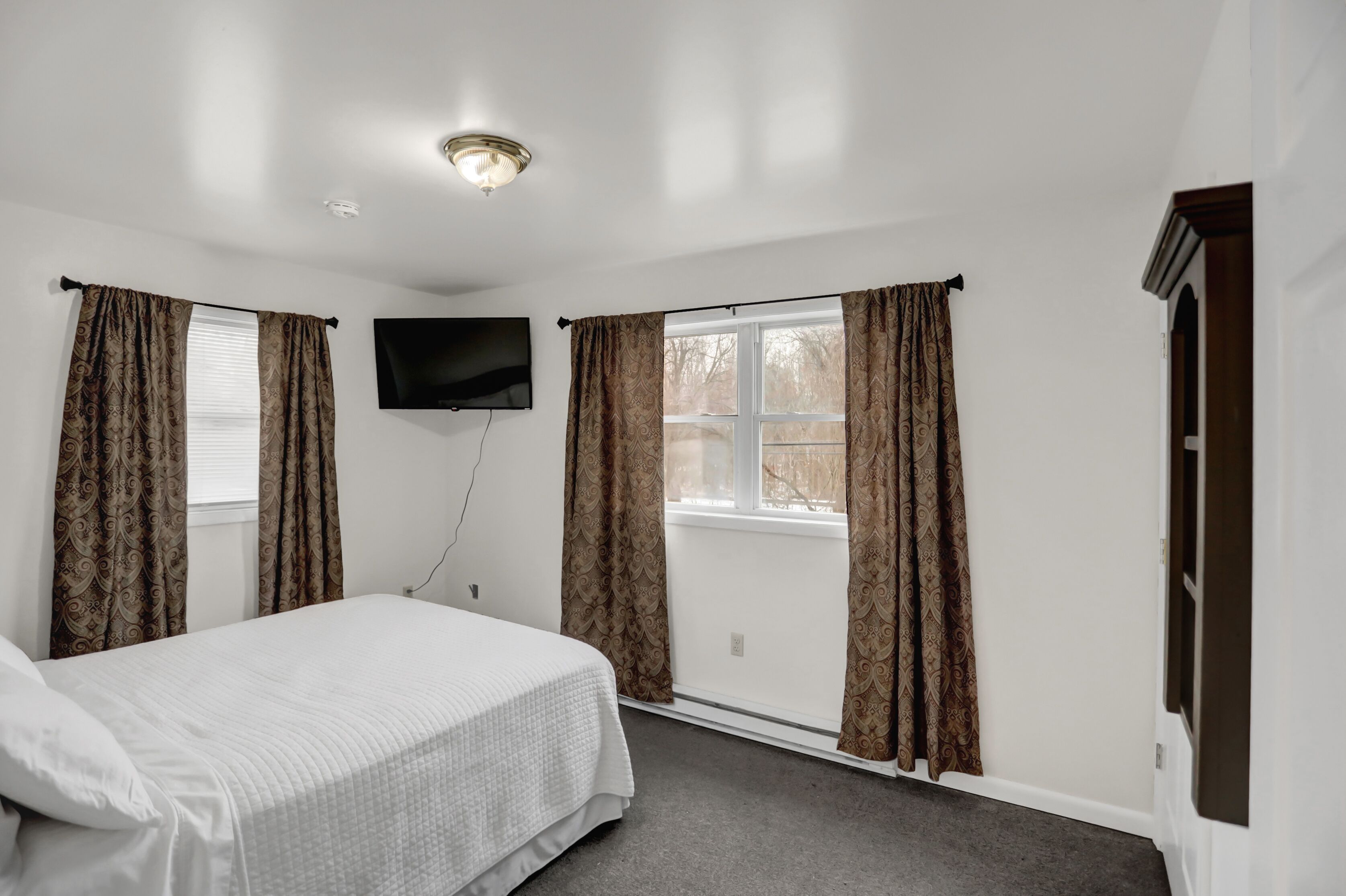 Un dormitorio con cama, dos ventanas con cortinas, un televisor montado en la pared y una lámpara de techo.