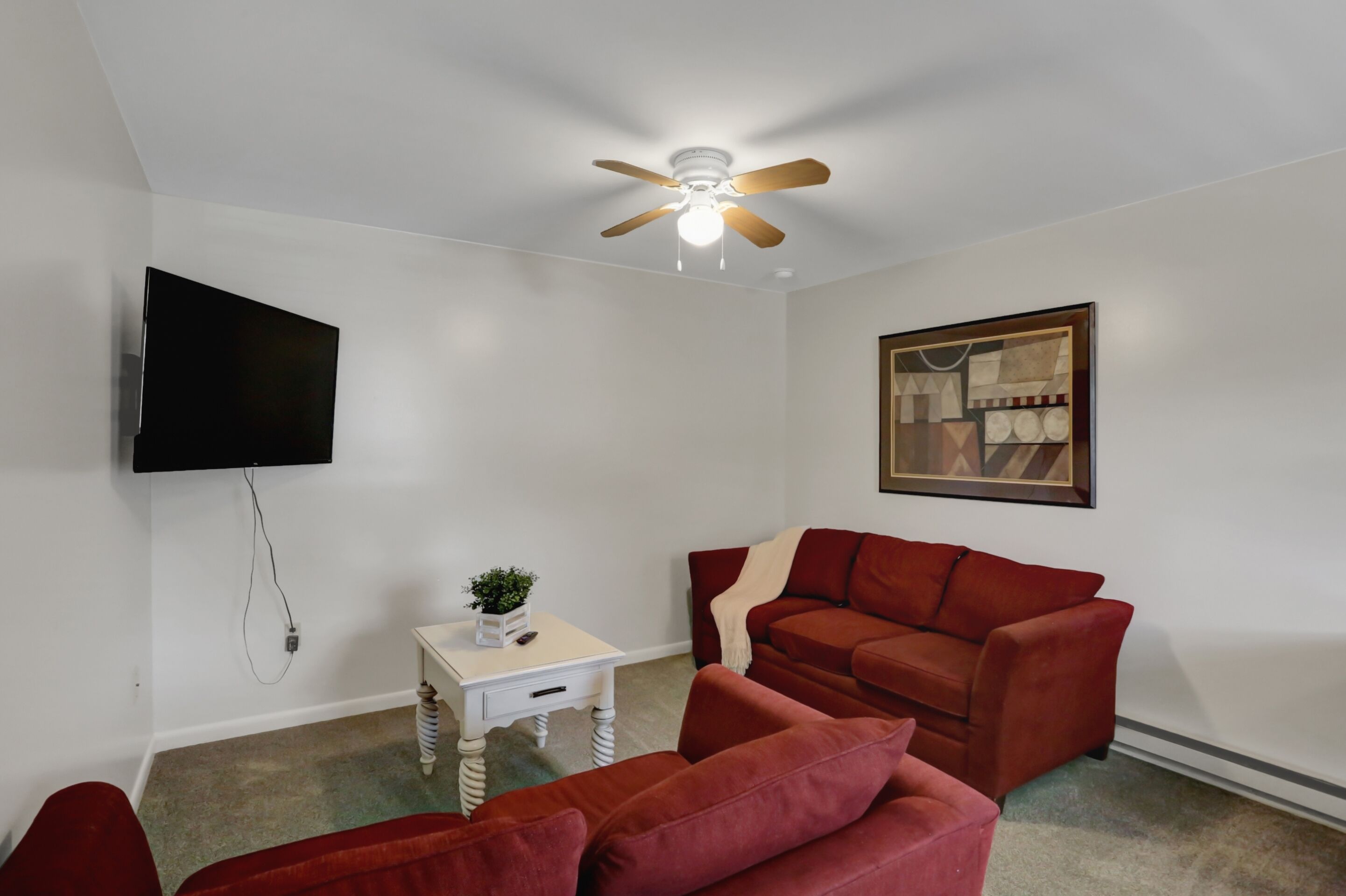 A living room with a red sofa, a white side table with a plant, a ceiling fan, a mounted TV, and a framed picture on the wall.