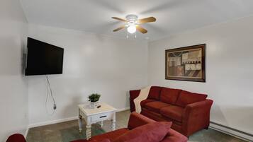 A living room with a red sofa, a white side table with a plant, a ceiling fan, a mounted TV, and a framed picture on the wall.