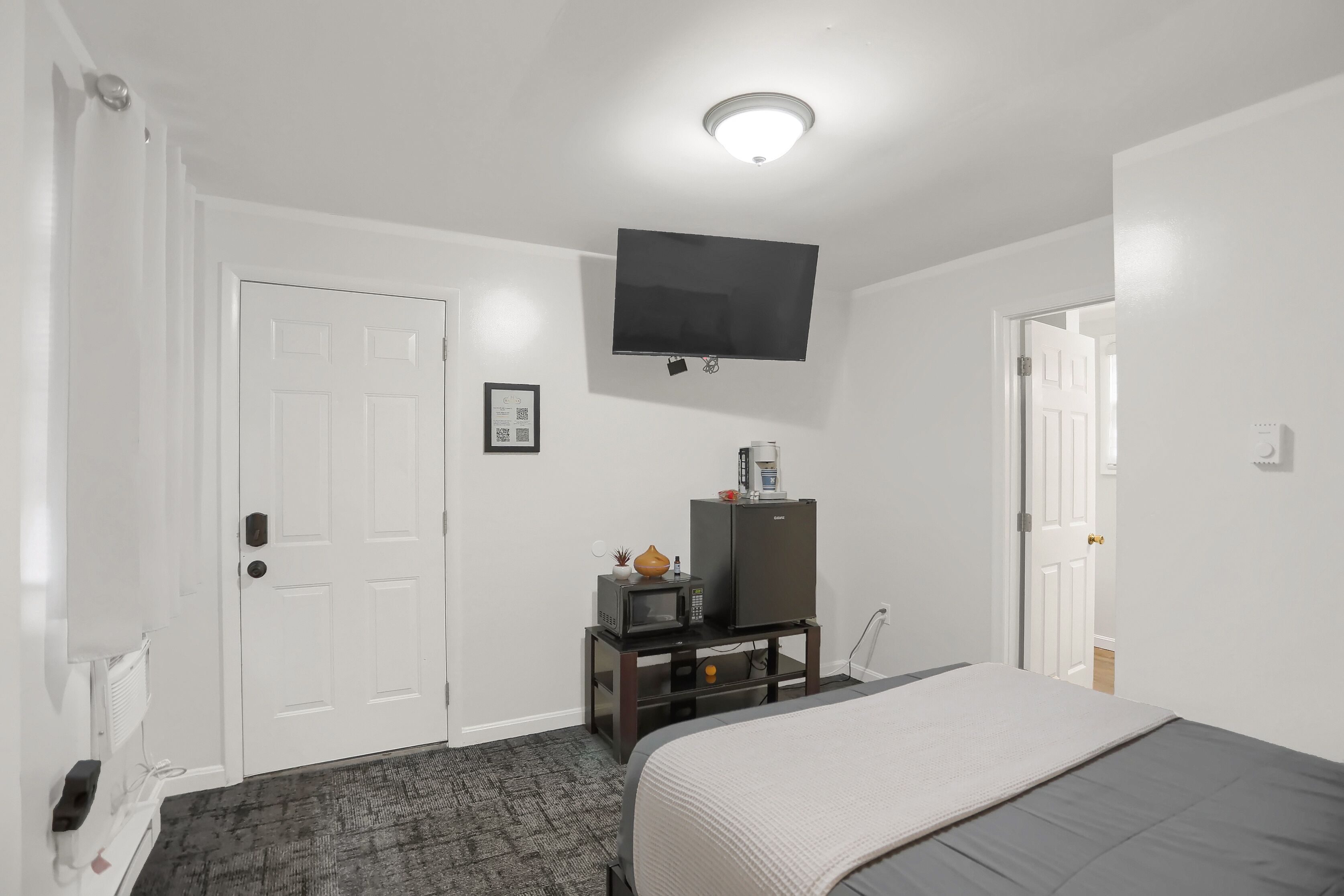 A hotel room with a bed, a TV mounted on the wall, a small table with a microwave and a refrigerator, and a door leading to another room.
