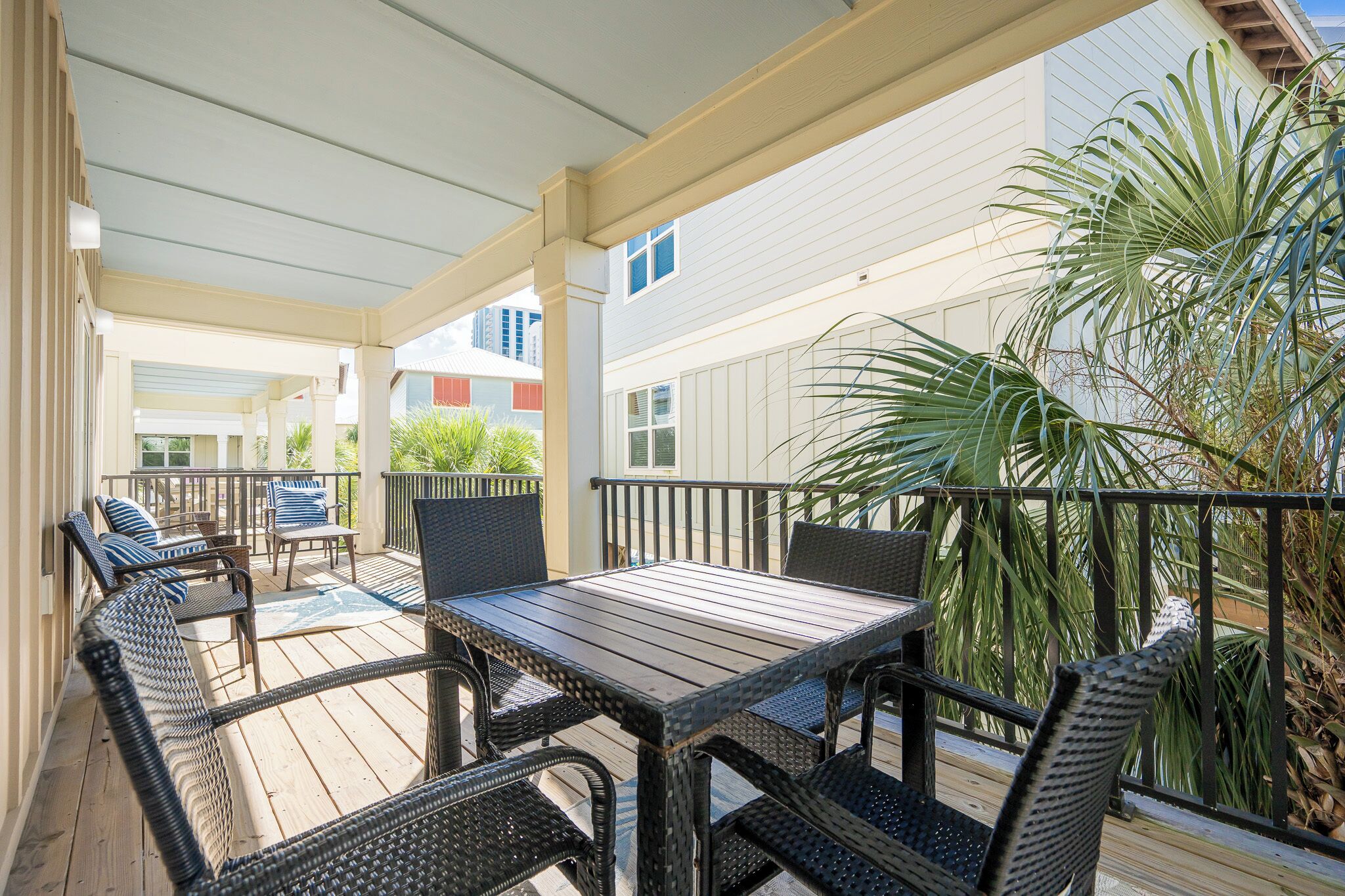 A covered outdoor patio with a wooden table and wicker chairs, surrounded by plants and a railing.