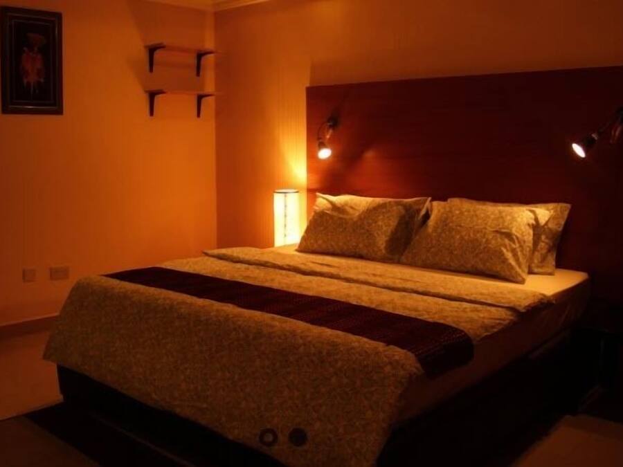 A bedroom with a bed, a nightstand, a wall-mounted lamp, and a potted plant.