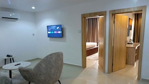 Quarto de hotel moderno com televisão de tela plana na parede, uma poltrona confortável, uma mesa redonda pequena e uma porta de madeira que dá para o banheiro.