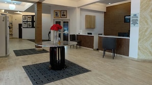 Uma área de recepção moderna com um balcão de mármore, uma mesa redonda com um vaso de flores vermelhas e um balcão de atendimento com painel de madeira ao fundo.