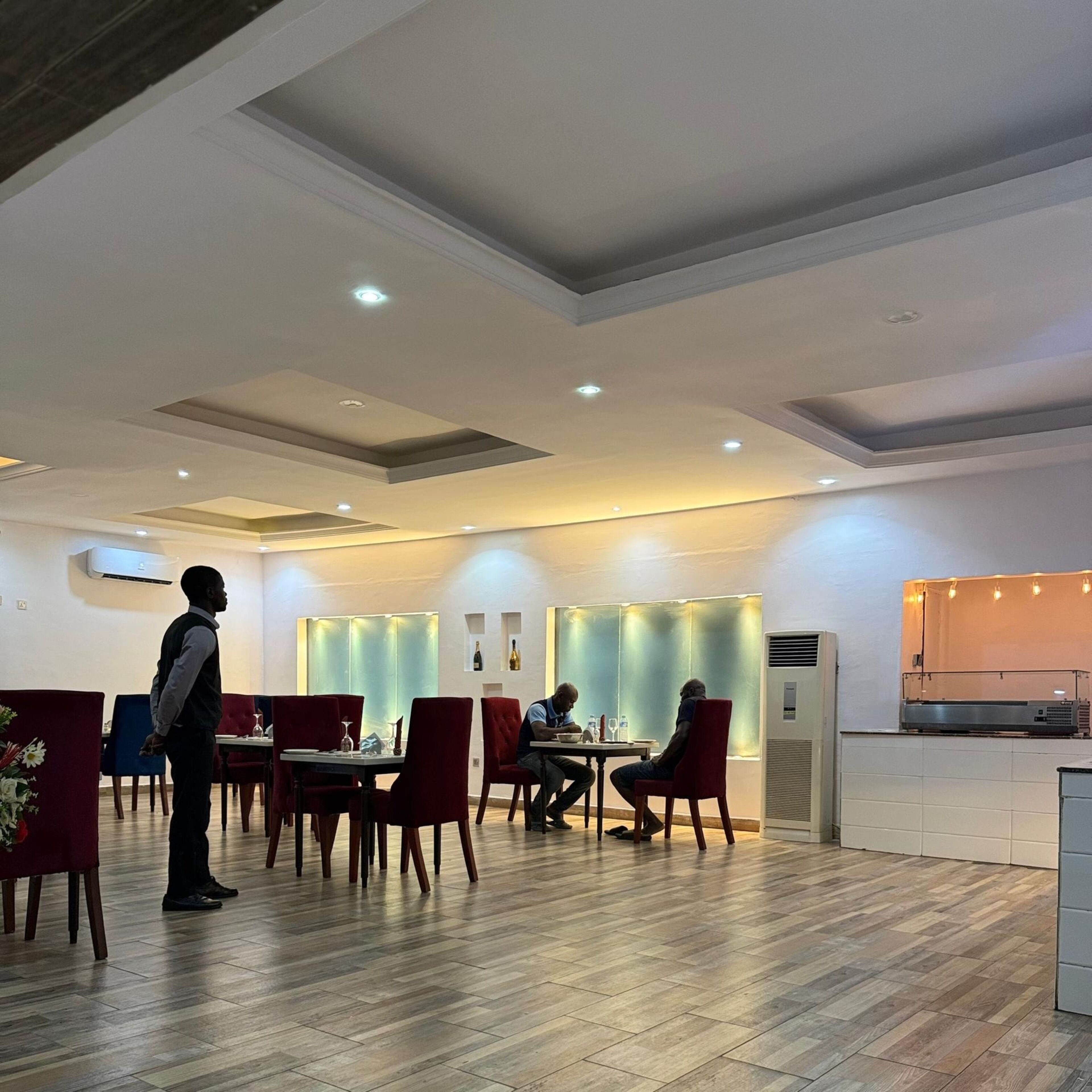 Ein Restaurant mit Holzboden, roten Stühlen und Tischen. Ein Kellner steht, zwei Gäste sitzen dort.