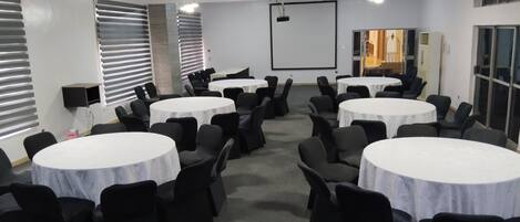 Un salón de conferencias con varias mesas redondas y sillas, una pantalla de proyector y ventanas amplias con persianas.