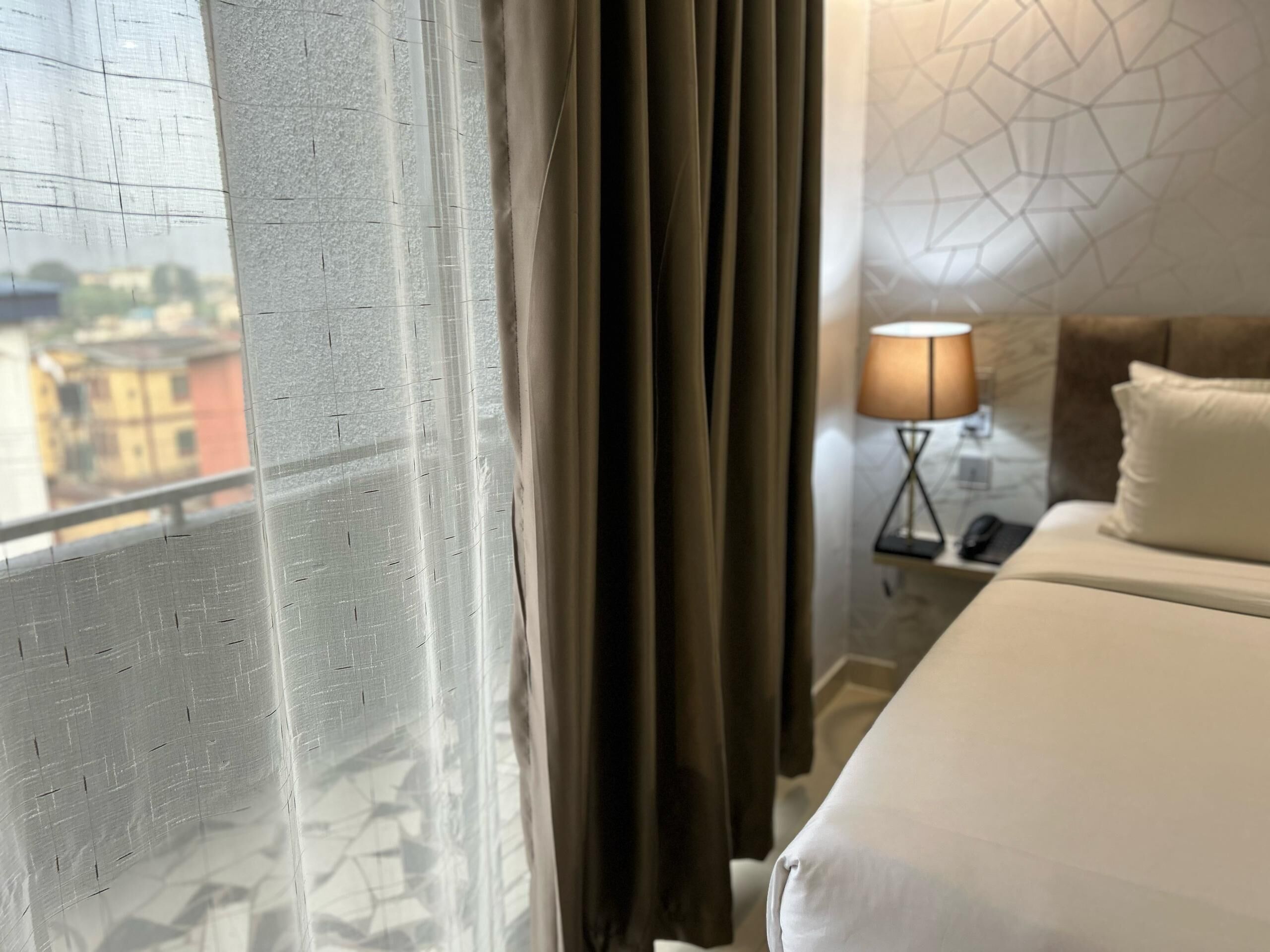 Quarto de hotel com cama, mesa de cabeceira, luminária e vista de prédios pela janela com cortinas.