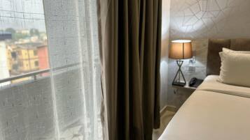 Quarto de hotel com cama, mesa de cabeceira, luminária e vista de prédios pela janela com cortinas.