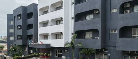 A multi-story building with balconies, air conditioning units, and a parking area with cars.