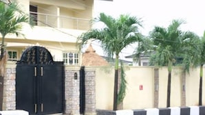 A two-story house with a black gate, a stone wall, and palm trees. - Class Suites Signature (Lagos)