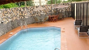A rectangular pool with a stone wall and a tiled deck. There are lounge chairs and a table with umbrellas. - Class Suites Signature (Lagos)